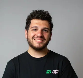 A young man with curly hair and a beard stands against a plain gray background, wearing a black Asphalt Green t-shirt and smiling at the camera.