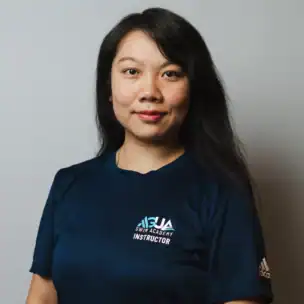 A woman with long dark hair wears a navy "Swim Academy Instructor" t-shirt and stands against a plain light background, facing the camera and smiling slightly.
