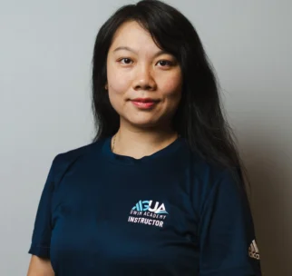 A woman with long dark hair wears a navy "Swim Academy Instructor" t-shirt and stands against a plain light background, facing the camera and smiling slightly.
