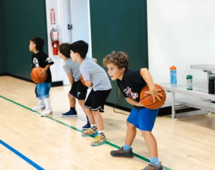 Four young boys in athletic clothes practice basketball drills indoors, each holding a basketball and bending their knees in a ready stance.