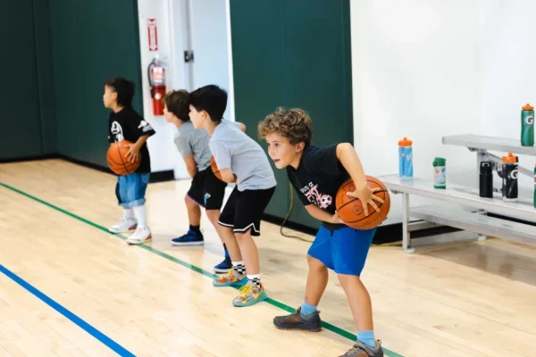 Four young boys in athletic clothes practice basketball drills indoors, each holding a basketball and bending their knees in a ready stance.