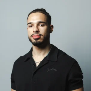 A man with short dark hair and a trimmed beard stands against a plain gray background, wearing a black collared shirt and a thin silver necklace.