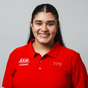 A person wearing a red lifeguard polo shirt with logos, smiling and standing against a plain light gray background.