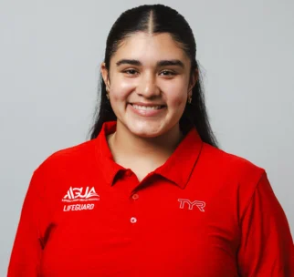A person wearing a red lifeguard polo shirt with logos, smiling and standing against a plain light gray background.