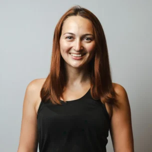 A woman with straight brown hair wearing a black sleeveless top smiles at the camera against a plain light gray background.
