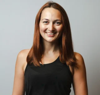 A woman with straight brown hair wearing a black sleeveless top smiles at the camera against a plain light gray background.