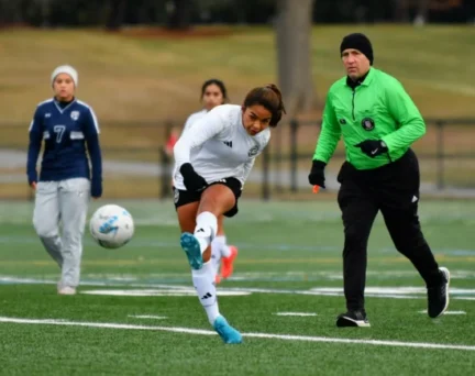 An Asphalt Green Soccer Club player in a white uniform kicks the ball during a match while a referee in green and other players look on.