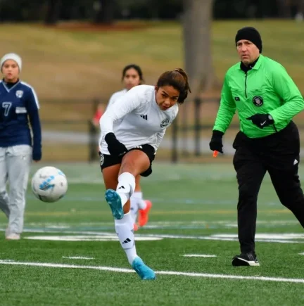 An Asphalt Green Soccer Club player in a white uniform kicks the ball during a match while a referee in green and other players look on.