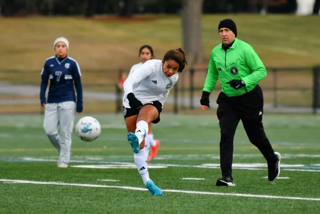 An Asphalt Green Soccer Club player in a white uniform kicks the ball during a match while a referee in green and other players look on.