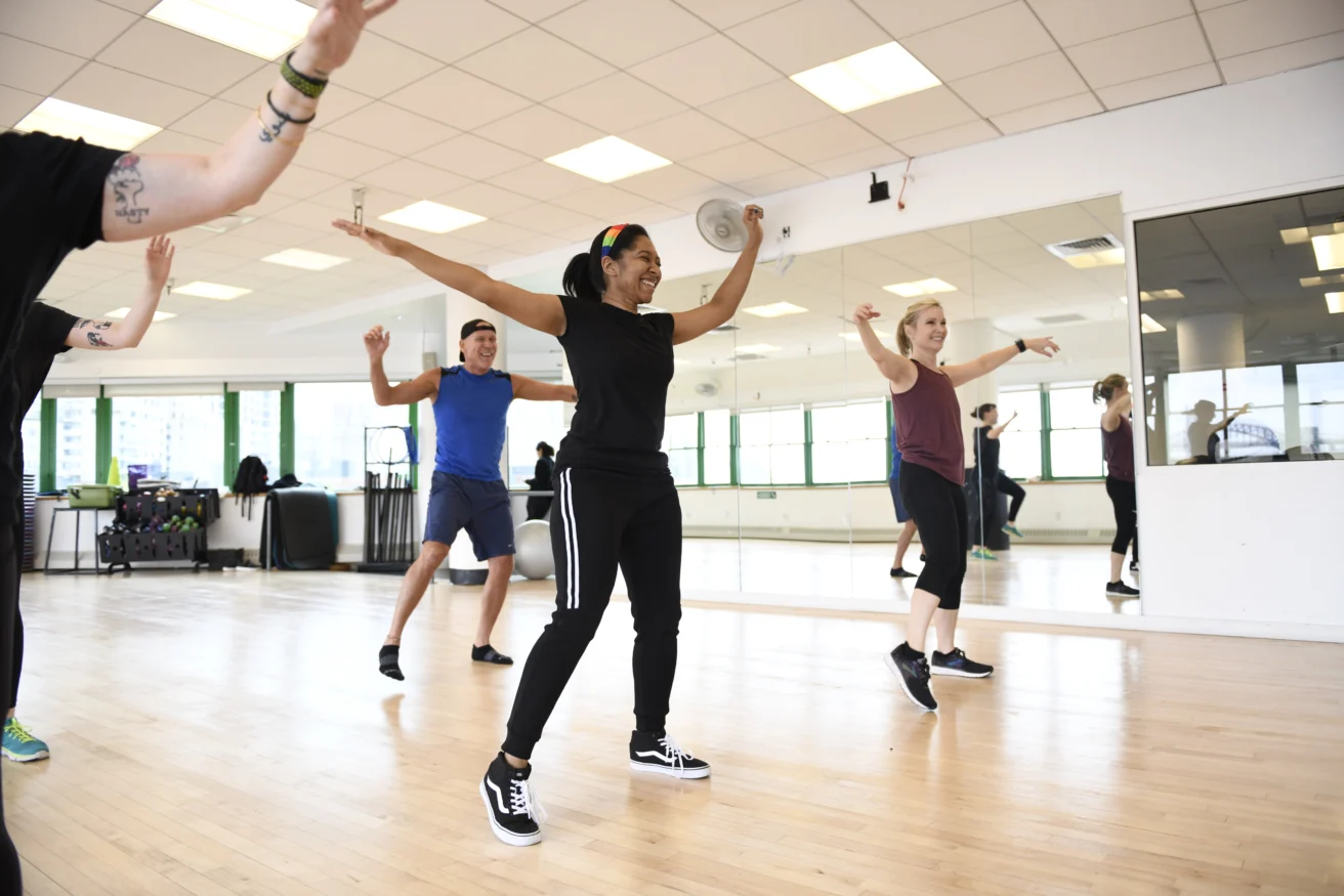 A group of people participate in a fitness dance class in a bright studio with wooden floors and mirrored walls.