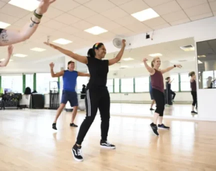 A group of people participate in a fitness dance class in a bright studio with wooden floors and mirrored walls.