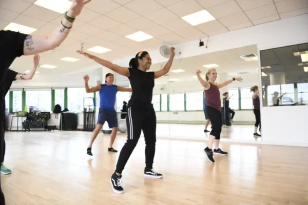 A group of people participate in a fitness dance class in a bright studio with wooden floors and mirrored walls.