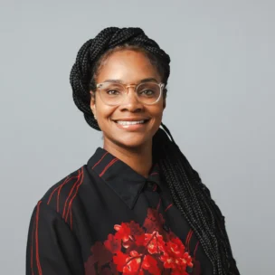 A woman with long braids, wearing glasses and a black shirt with red floral patterns, smiles at the camera against a plain gray background.