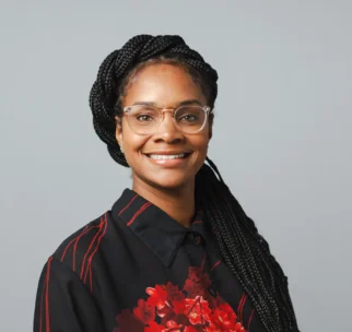 A woman with long braids, wearing glasses and a black shirt with red floral patterns, smiles at the camera against a plain gray background.