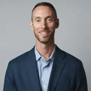 A man wearing a blue suit jacket and light blue shirt stands in front of a plain, light gray background, smiling at the camera.