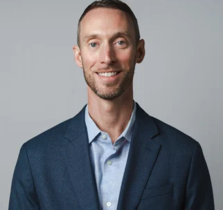 A man wearing a blue suit jacket and light blue shirt stands in front of a plain, light gray background, smiling at the camera.