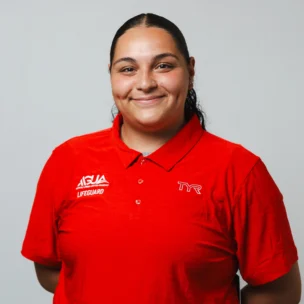 A person with long dark hair, wearing a red lifeguard uniform shirt with "TYR" and "AQUA LIFEGUARD" logos, stands against a plain light background and smiles at the camera.