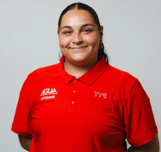 A person with long dark hair, wearing a red lifeguard uniform shirt with "TYR" and "AQUA LIFEGUARD" logos, stands against a plain light background and smiles at the camera.