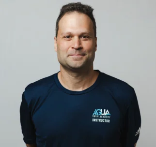 A man wearing a navy blue "Swim Academy Instructor" t-shirt stands against a plain light background, facing the camera with his hands behind his back.