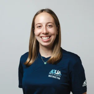 A woman with straight, shoulder-length hair smiles at the camera, wearing a navy blue "SJA Swim Academy Instructor" shirt and a necklace, standing against a plain background.
