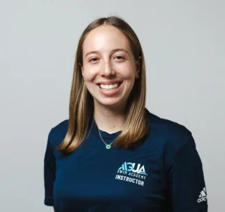 A woman with straight, shoulder-length hair smiles at the camera, wearing a navy blue "SJA Swim Academy Instructor" shirt and a necklace, standing against a plain background.