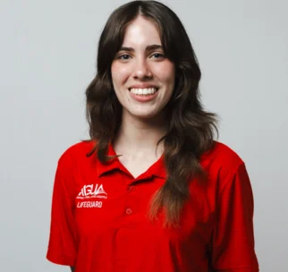 A person with long brown hair wearing a red lifeguard polo shirt smiles at the camera against a plain light background.