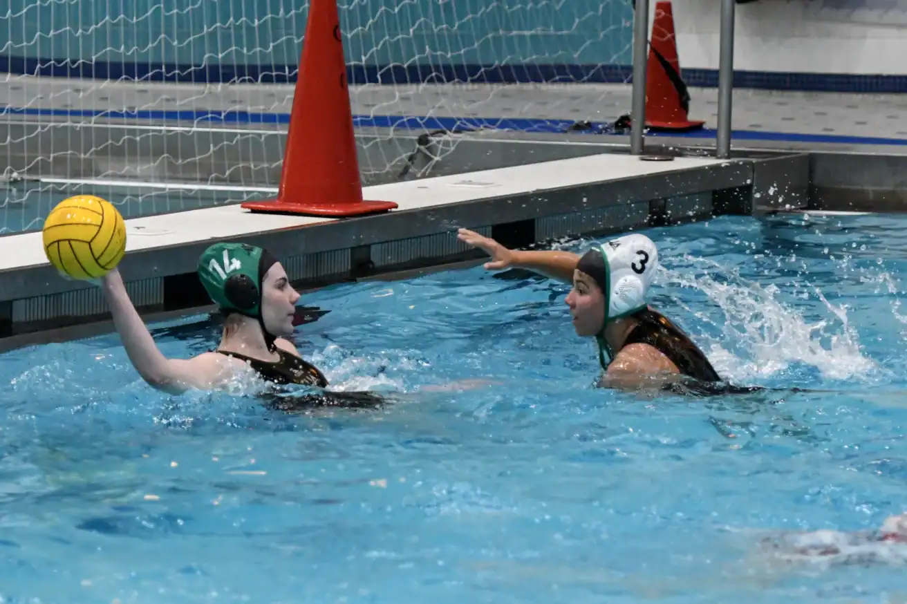 Two female water polo players in swim caps compete in a pool; one holds a yellow ball while the other defends, with a goal and orange cones in the background.