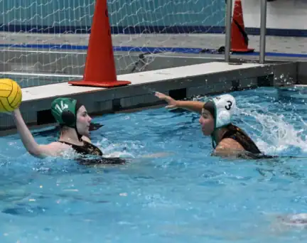 Two female water polo players in swim caps compete in a pool; one holds a yellow ball while the other defends, with a goal and orange cones in the background.