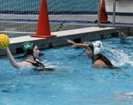 Two female water polo players in swim caps compete in a pool; one holds a yellow ball while the other defends, with a goal and orange cones in the background.