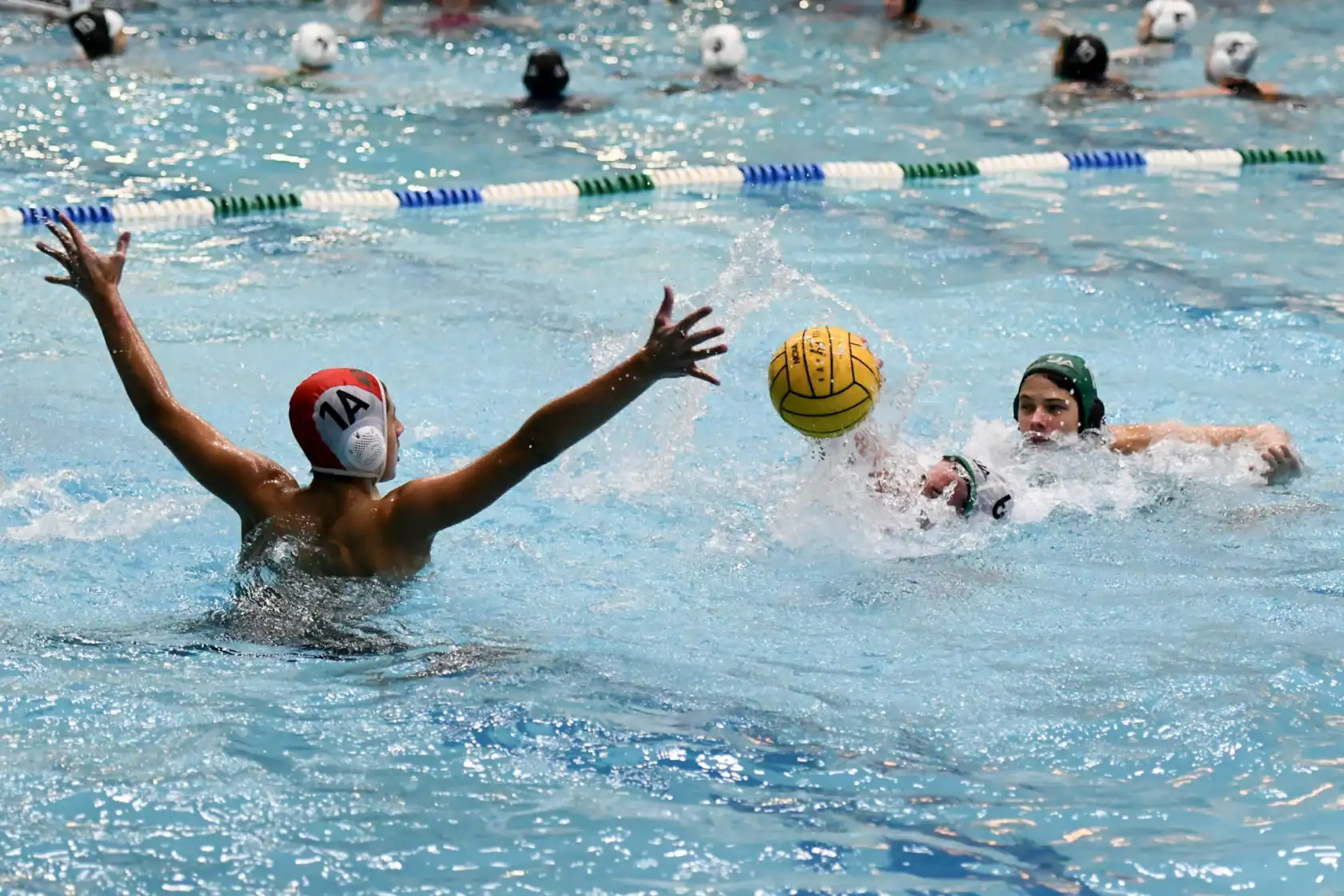 Two water polo players compete for the ball in a pool, with one player attempting a block and others swimming in the background.