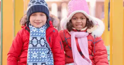 Two children in warm winter clothes and hats sit side by side outdoors, smiling at the camera.