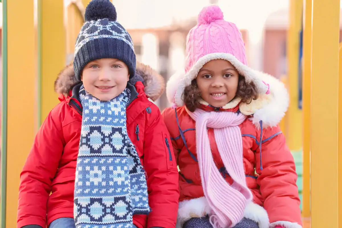 Two children in warm winter clothes and hats sit side by side outdoors, smiling at the camera.