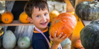 A young boy smiles while holding a large orange pumpkin, surrounded by various pumpkins and gourds on shelves.