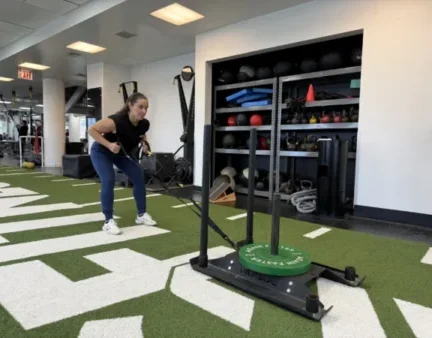 Asphalt Green Director of Fitness Nicole Lee pulls a weighted sled with resistance straps across artificial turf inside a gym, with various equipment visible in the background.