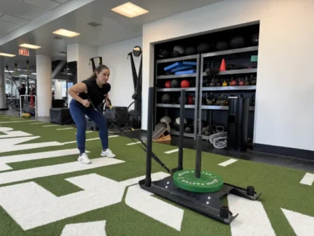 Asphalt Green Director of Fitness Nicole Lee pulls a weighted sled with resistance straps across artificial turf inside a gym, with various equipment visible in the background.