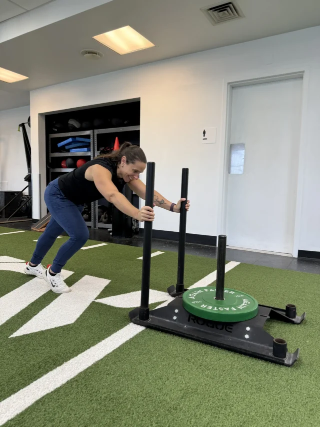 Person pushes a weighted sled on an indoor turf track in a gym setting, demonstrating a strength and conditioning exercise.