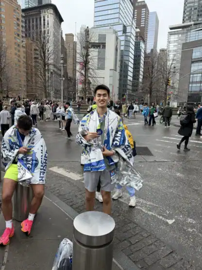 A runner wearing a race bib and a finisher's wrap smiles and holds a medal on a city street during the NYC Marathon, with other runners and tall buildings in the background.