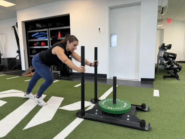 A woman in athletic wear pushes a weighted sled on artificial turf inside a gym, with workout equipment visible in the background.