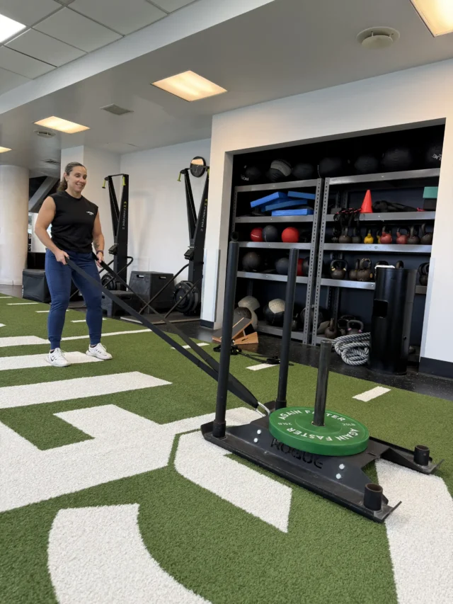 A person pulls a weighted sled across artificial turf in a gym, with exercise equipment and medicine balls in the background.