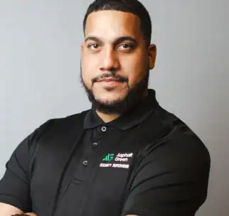 A man with short hair and a beard, wearing a black "Asphalt Green Security Supervisor" polo shirt, stands with his arms crossed against a plain gray background.