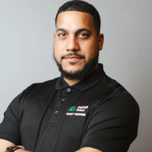 A man with short hair and a beard, wearing a black "Asphalt Green Security Supervisor" polo shirt, stands with his arms crossed against a plain gray background.