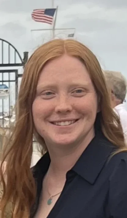 A person with long red hair smiles outdoors near a black metal fence, with an American flag and cloudy sky in the background.