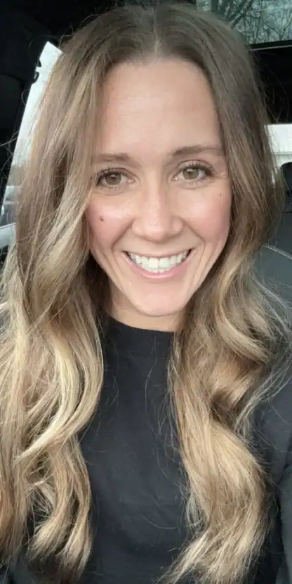 A woman with long, wavy brown hair and a black sweater smiles at the camera while sitting in a car.