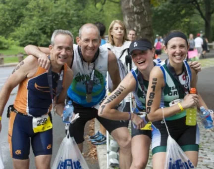 Four triathletes wearing race numbers and medals pose and smile, holding bags and drinks, reminiscent of NYC Marathon finishers, with a park setting and other people in the background.