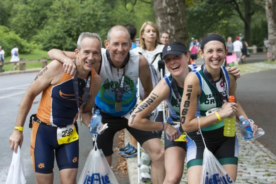 Four triathletes wearing race numbers and medals pose and smile, holding bags and drinks, reminiscent of NYC Marathon finishers, with a park setting and other people in the background.