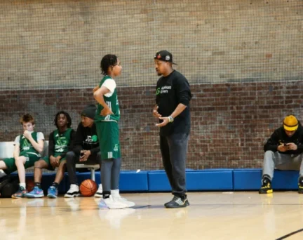 A youth basketball coach talks to a player on the court while teammates sit on the bench and another person uses a phone nearby.