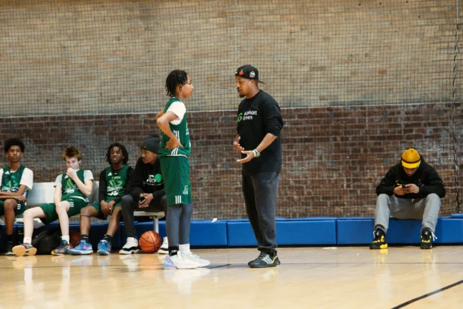 A youth basketball coach talks to a player on the court while teammates sit on the bench and another person uses a phone nearby.