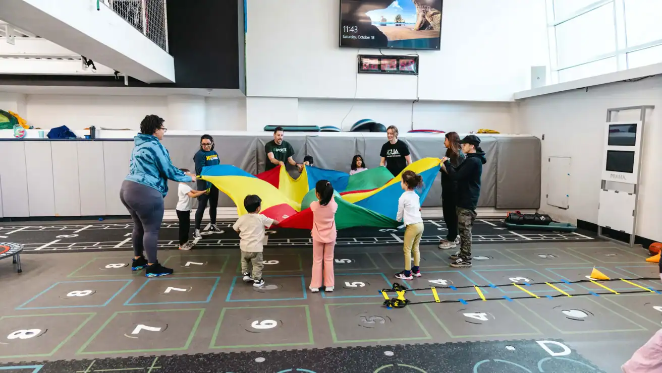 Children and adults stand in a gymnasium holding a colorful parachute together, engaging in a group activity. Various sports equipment is visible in the background.