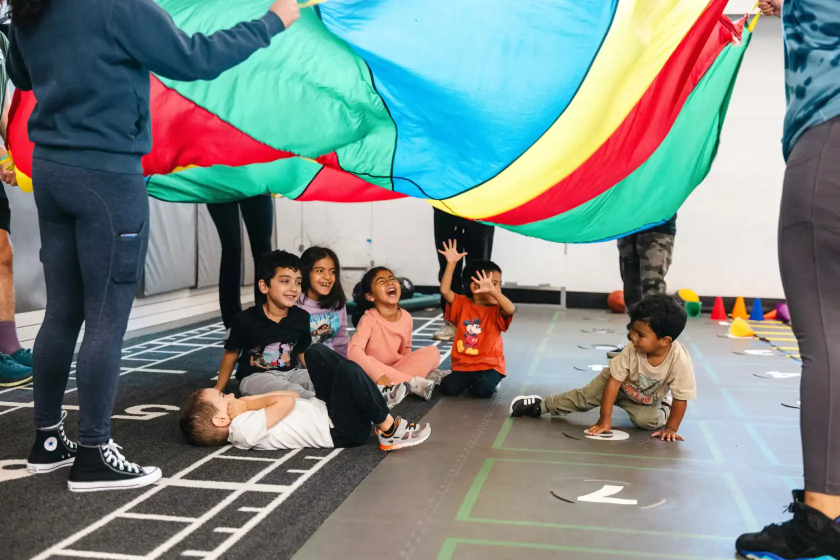 A group of children sit and play under a large colorful parachute held by adults in an indoor gym setting.