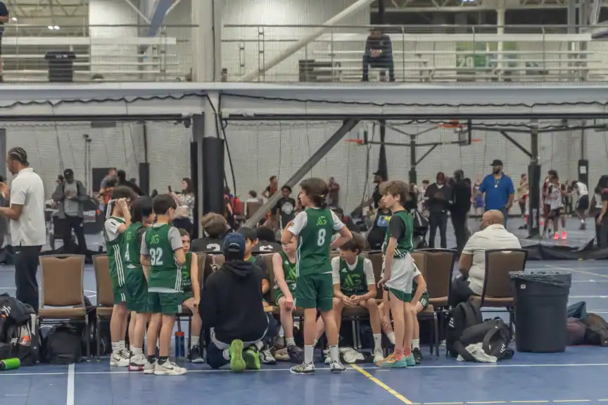 An AG Basketball NYC youth basketball team in green uniforms huddles around their coach on an indoor court, discussing basketball skills, while other games and people are visible in the background.